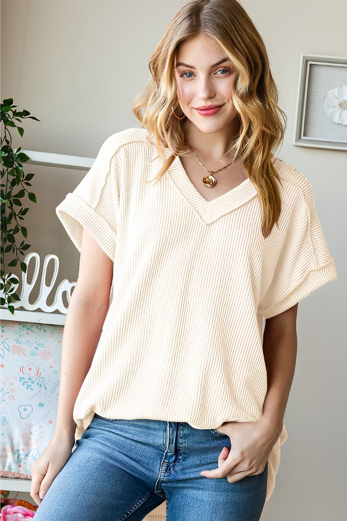 A young woman with wavy blonde hair smiles indoors, wearing the Solid Ribbed Dolman Top in cream and blue jeans. She poses with one hand in her pocket, surrounded by plants and decor that reflect Heimish USA’s effortless style.