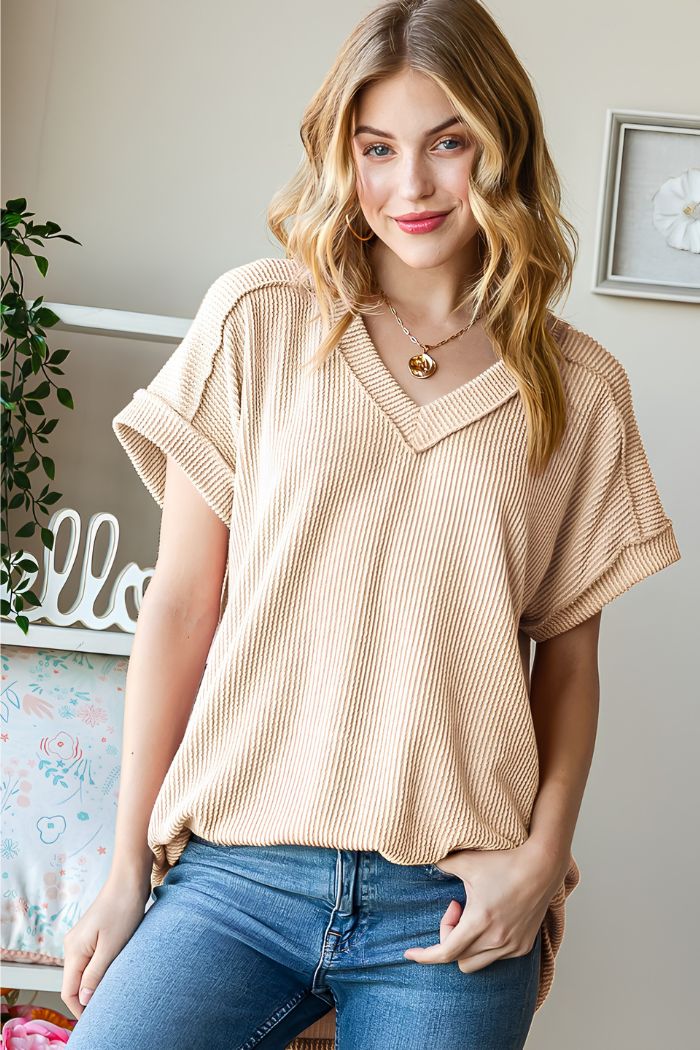 A young woman with wavy blonde hair smiles at the camera indoors, wearing the Solid Ribbed Dolman Top in beige and blue jeans. A plant, shelf, and decorative pillow in the background create a cozy Heimish USA vibe.