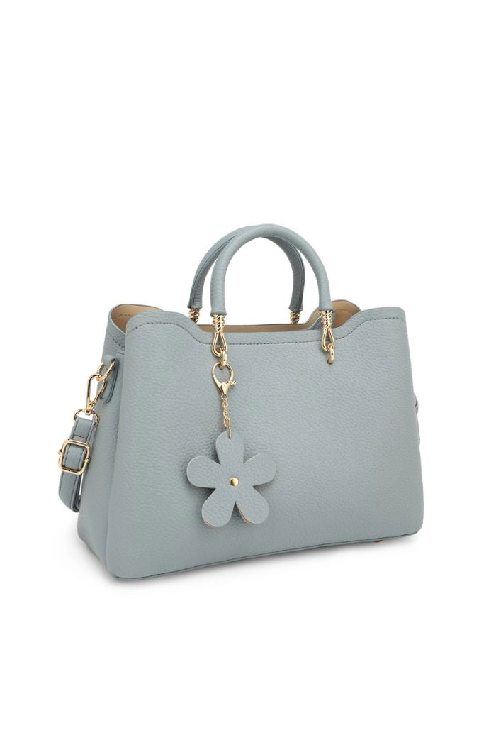 The Fatima Flower Satchel is a light blue faux leather bag featuring gold hardware, double handles, a detachable shoulder strap, and a decorative flower-shaped charm on the front for a stylish, structured look.
