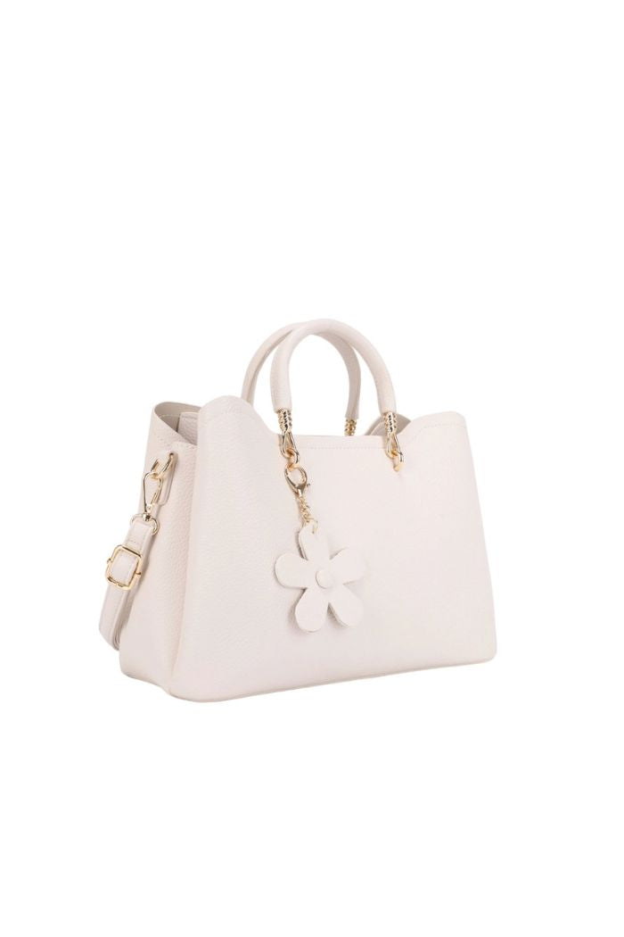 The Fatima Flower Satchel is a white faux leather bag with gold hardware, top handles, a detachable shoulder strap, and a flower-shaped charm on one handle, shown on a white background.