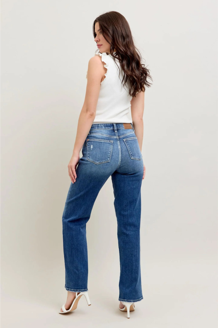 A woman viewed from the back, wearing dark blue, high-waisted, straight-leg jeans with slight distressing on the pockets, a white sleeveless top with a ruffled edge, and white high-heeled sandals. She has long, dark, wavy hair and is looking over her right shoulder.