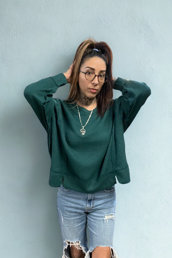 A woman with a high ponytail and glasses, wearing a dark green, oversized, round neck waffle knit long-sleeved top, ripped blue jeans, and a silver necklace, stands with her hands behind her head against a light blue textured wall.