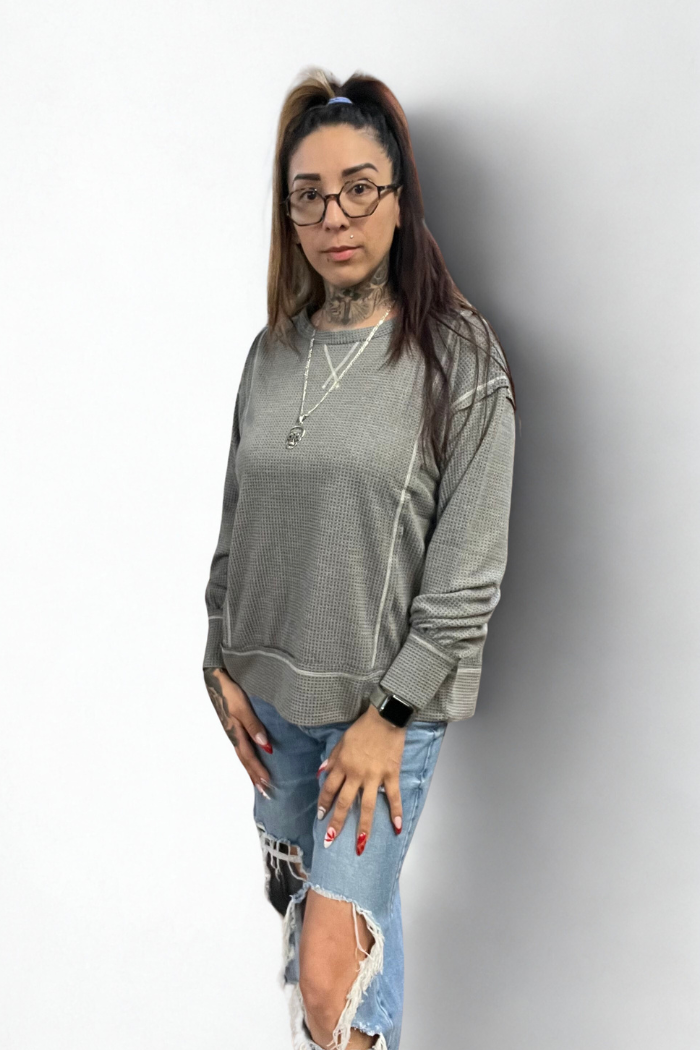 A person with glasses, long brown and blonde hair in a ponytail, wearing a grey waffle-knit long-sleeve shirt with contrast stitching, distressed blue jeans, a silver necklace, and visible neck and hand tattoos, stands in front of a white wall.
