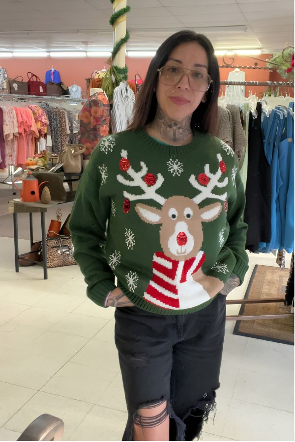 A woman with glasses and tattoos stands in a boutique with clothing and bags displayed in the background, wearing the Sequin Reindeer Christmas Sweater paired with black ripped jeans. Her festive sweater adds a cheerful holiday touch to the stylish atmosphere.