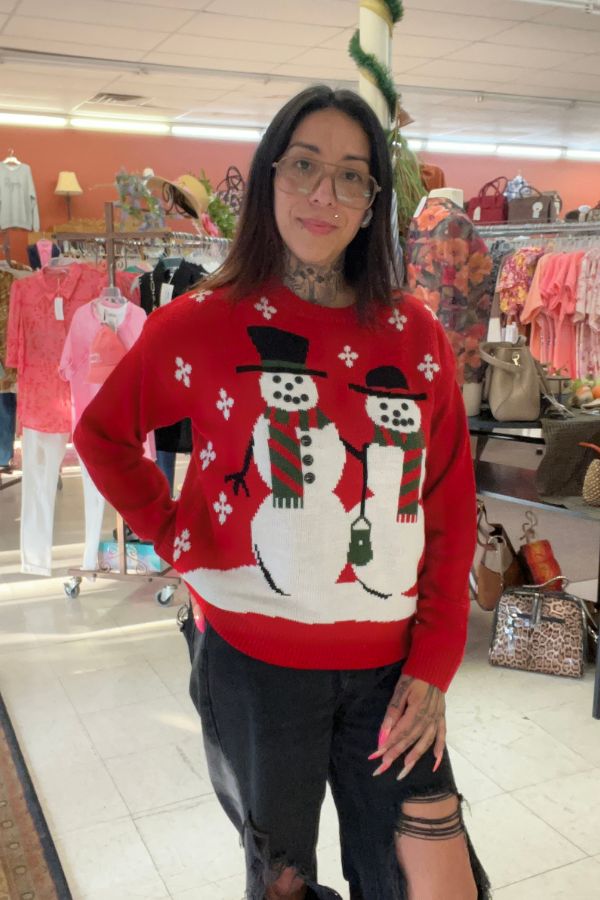 A woman with glasses and tattoos wears the Snowman Christmas Knit Sweater, featuring two cheerful snowmen, while standing in a clothing store filled with colorful shirts, handbags, and assorted accessories.
