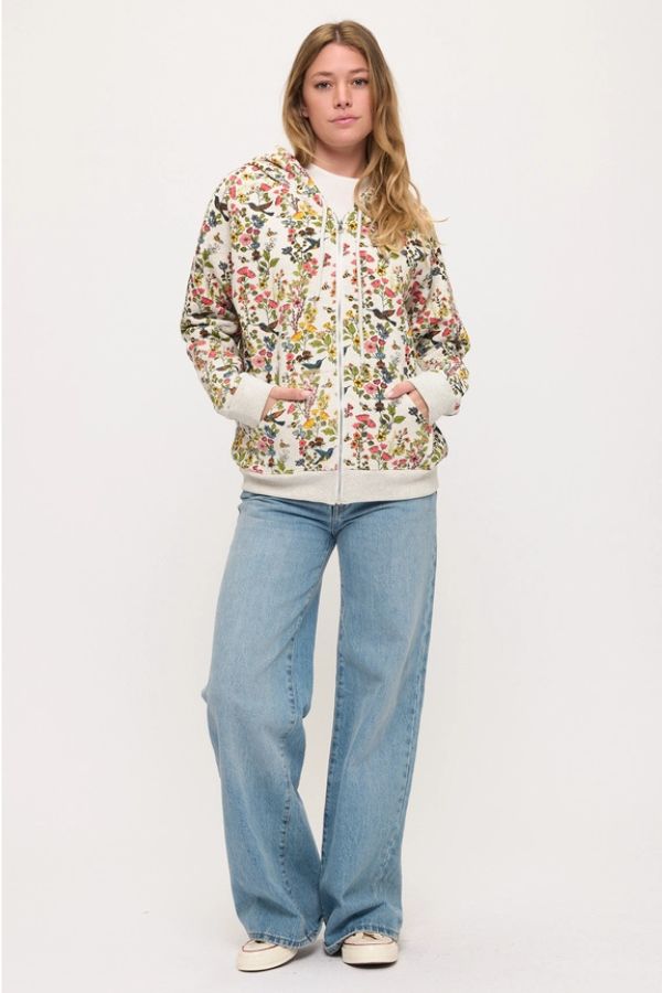 A woman stands against a white background, wearing the Floral Hummingbird Zip Hoodie with a relaxed fit, paired with light blue wide-leg jeans and white sneakers. She has long, wavy hair and a neutral expression.