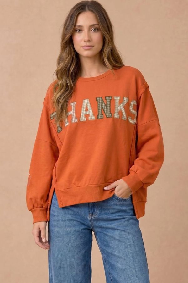 A woman wears the Plaid Thanks Patchwork Sweatshirt in an oversized fit with plaid lettering, paired with loose blue jeans, standing against a beige background, hand in her pocket.
