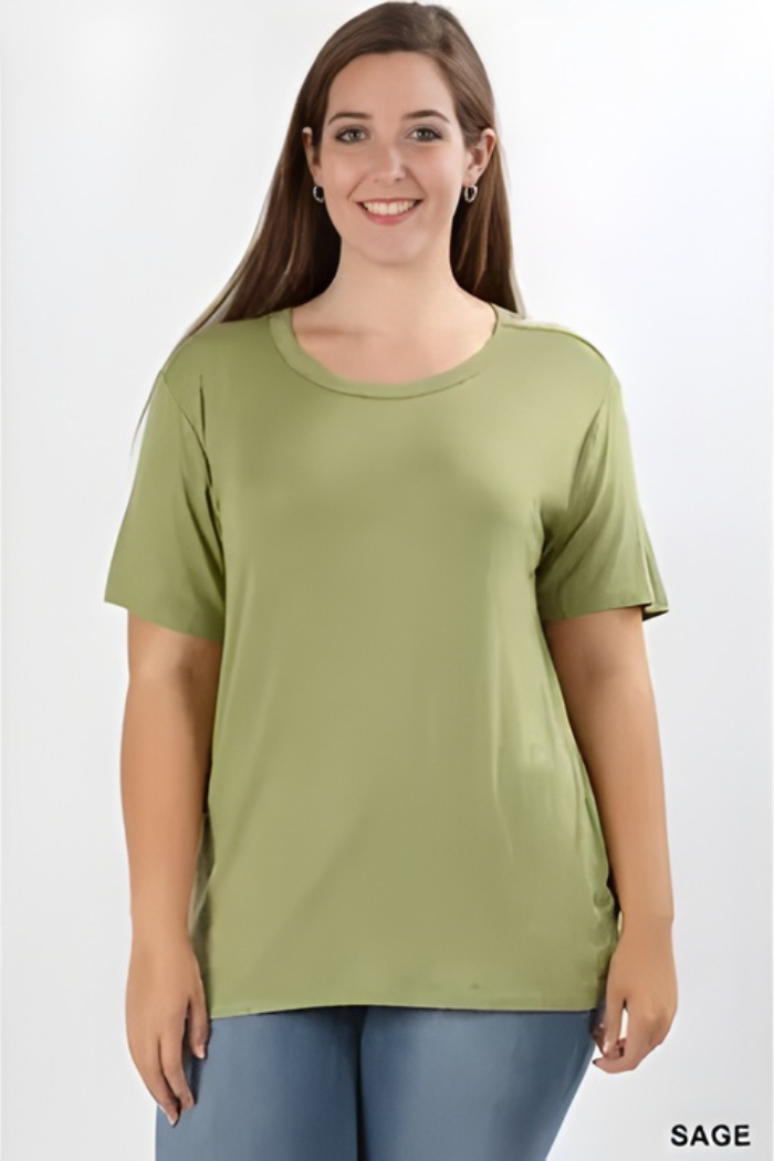 A woman with long brown hair smiles while wearing a loose-fitting sage green Plus Size Rayon Tee and blue jeans, standing against a plain white background.