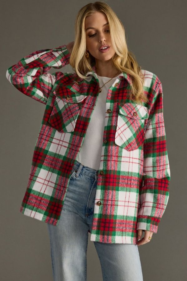 A woman wearing the Holiday Plaid Cozy Shacket—a red, green, and white plaid flannel shirt with a button-front closure—layered over a white top and blue jeans, stands against a gray background with one hand touching her hair, making for a perfect cozy fall look.