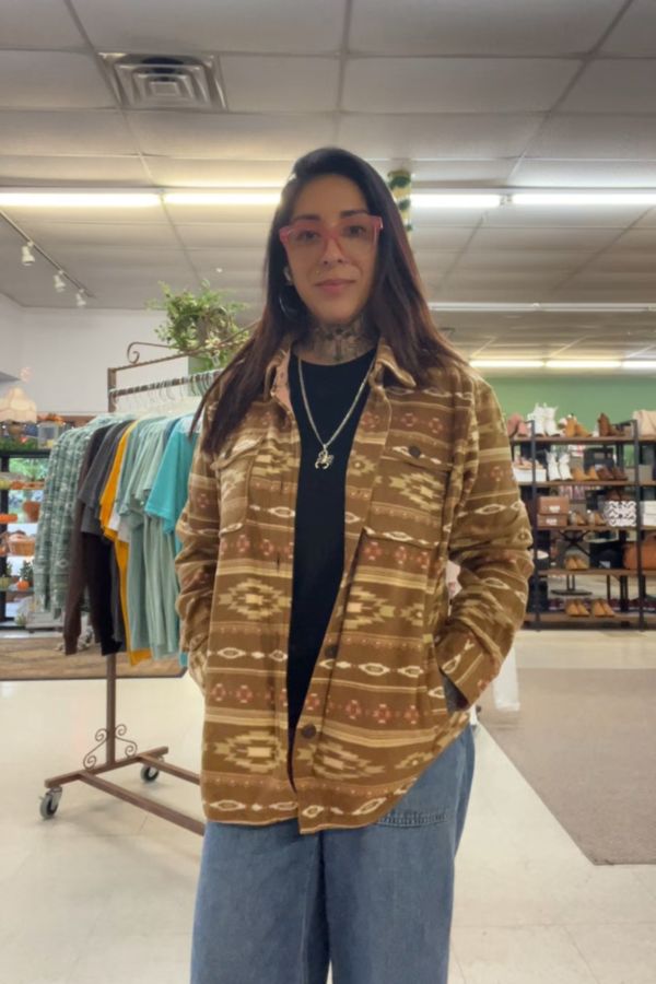 A woman wearing a Southwestern Fleece Shacket, black shirt, jeans, and red glasses stands inside a clothing store with racks of clothes and shoes in the background.