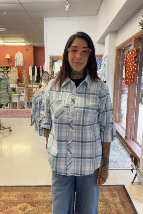 A person with tattoos, glasses, and long dark hair is wearing the Plaid Button Shirt and blue jeans, standing in a clothing store filled with racks of clothes and warm lighting in the background.