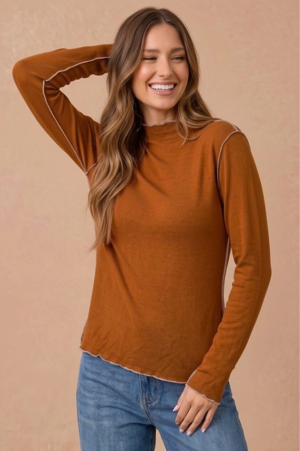 A woman wearing a Curvy Frilled Mock Neck Hacci Top in rust, featuring a frilled mock neckline, exposed light-colored seam detailing, and a ruffled hem.