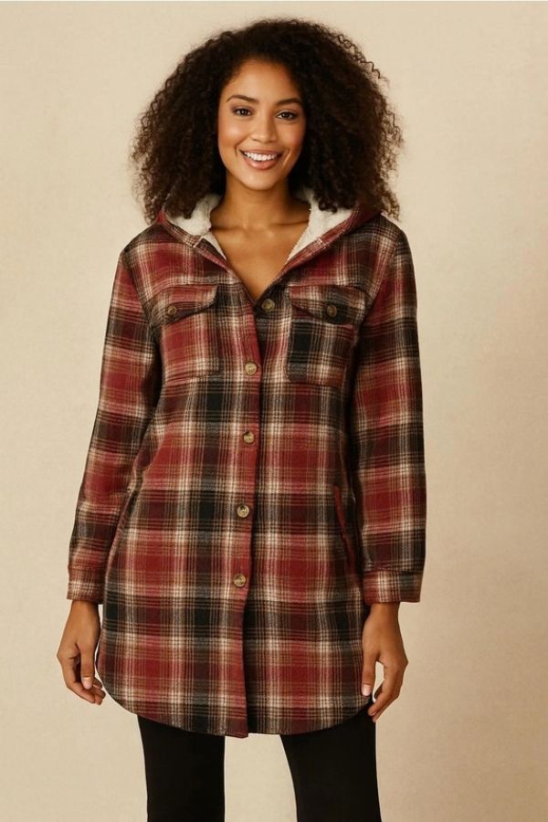 A woman with curly hair smiles while wearing the Fur Lined Plaid Hooded Shacket, a long, button-up red and brown plaid jacket with a hood and two chest pockets.