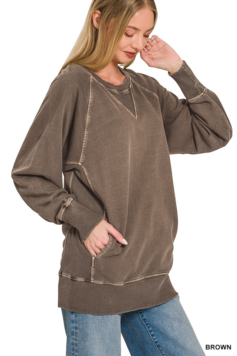 A woman stands against a plain white background, wearing the Pigment Dyed Pullover—a relaxed fit, brown French terry top with visible seams—paired with blue jeans. One hand is in her pocket while the other touches her face.