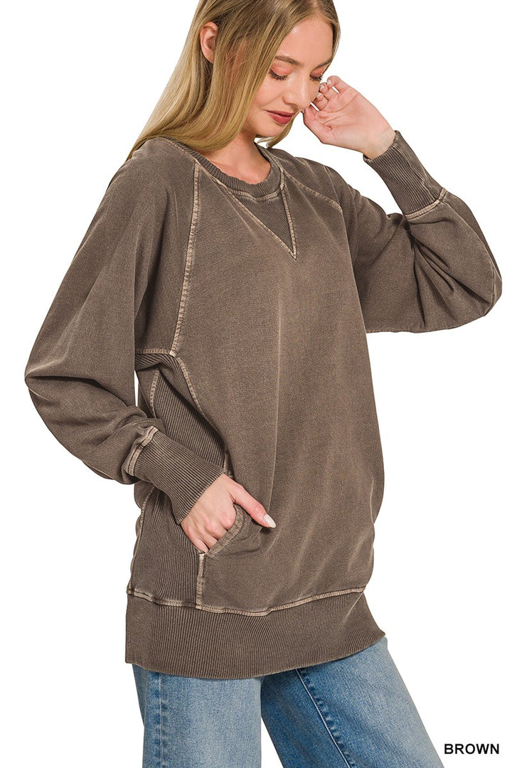 A woman stands against a plain white background, wearing the Pigment Dyed Pullover—a relaxed fit, brown French terry top with visible seams—paired with blue jeans. One hand is in her pocket while the other touches her face.