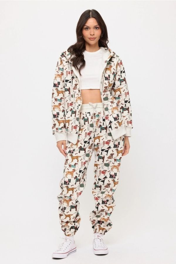 Wearing the Holiday Dogs Zip Up Hoodie and matching pants set, featuring a colorful all-over dog print along with a white crop top and sneakers, a woman poses against a plain white background—an ideal look for dog lovers.