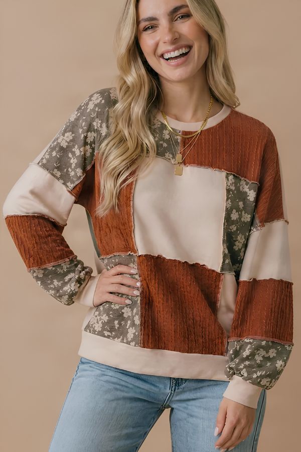 A smiling woman wears the Color Block Knit Top with rust, cream, and floral patterns in a relaxed fit, paired with light blue jeans, standing against a beige background.