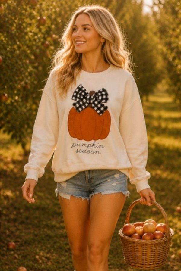 A woman with long blonde hair strolls through an orchard on a sunny day, wearing the Pumpkin Season Bow Sweatshirt and denim shorts, holding a basket of apples.
