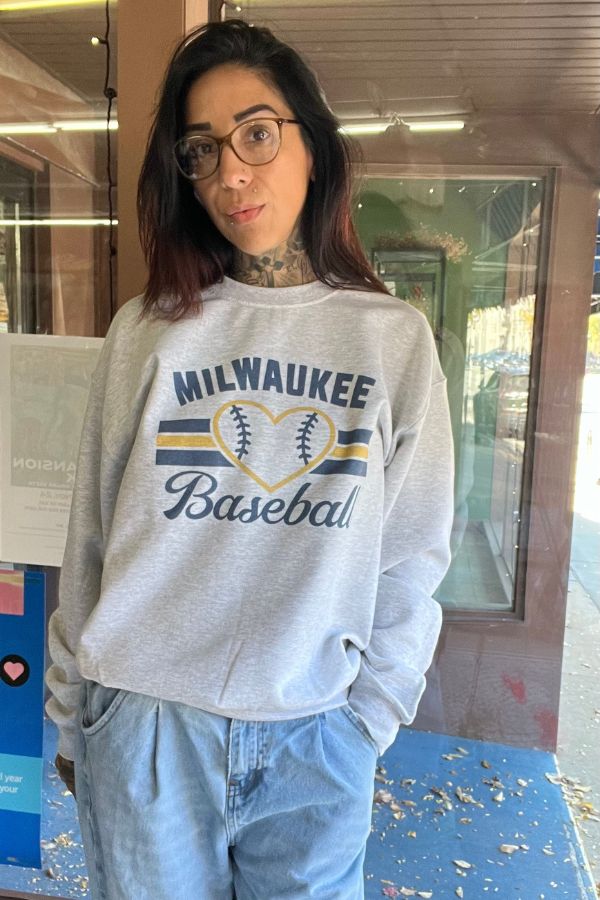 A person wears a light gray Milwaukee Baseball Crewneck Sweatshirt featuring blue text and a yellow heart outlined with baseball stitching in the center.