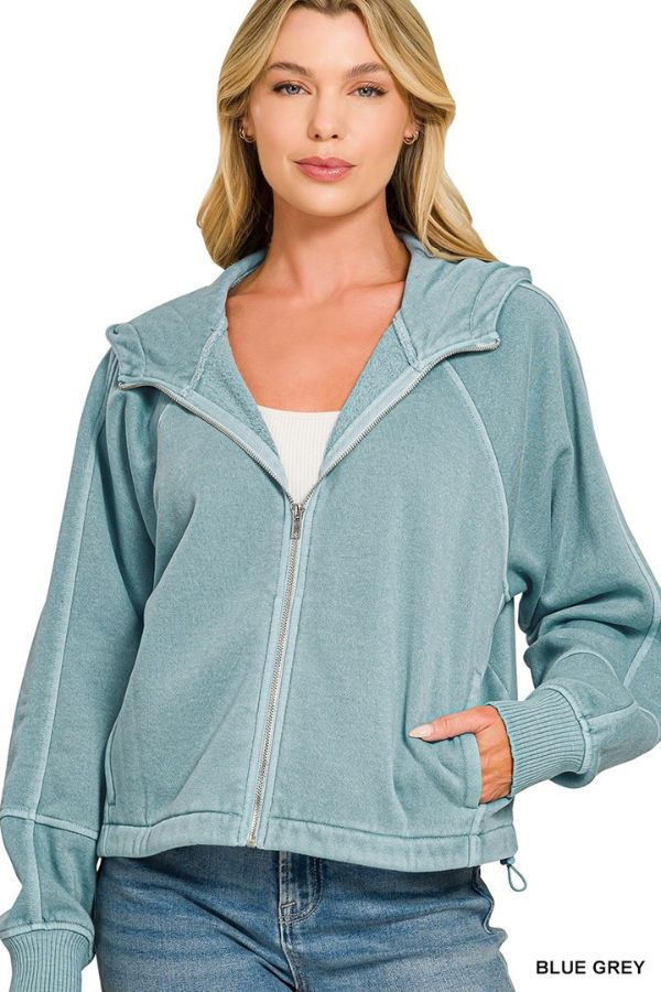 Woman wearing the Pigment Dye Zip Up Hoodie in light blue-grey with front pockets and a white top underneath, posing with one hand in her pocket. “BLUE GREY” text shown at bottom right.