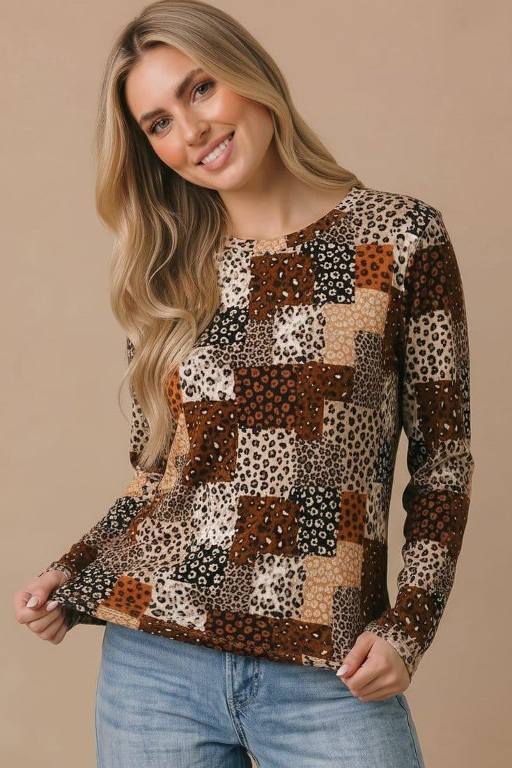 A woman with long blonde hair wears the Animal Patchwork Top—a relaxed fit, long sleeve blouse with a patchwork design featuring various animal prints in brown, tan, and black tones—paired with light blue jeans. She stands against a beige background and smiles.
