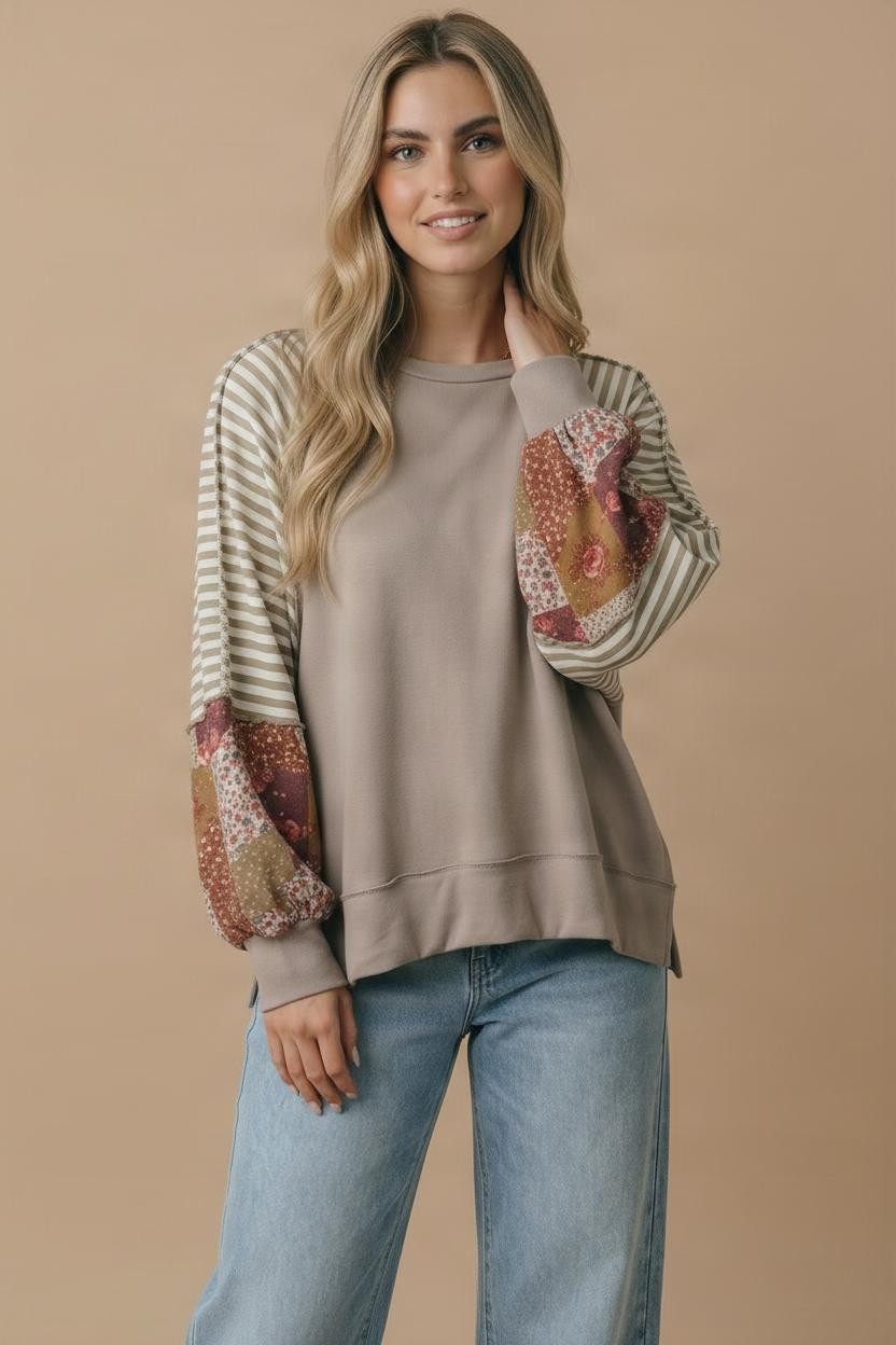 A woman with long, wavy blonde hair smiles gently at the camera, standing against a beige background. She wears the Patchwork Bubble Sleeve Top in taupe French terry with mixed print sleeves, paired with light blue jeans.
