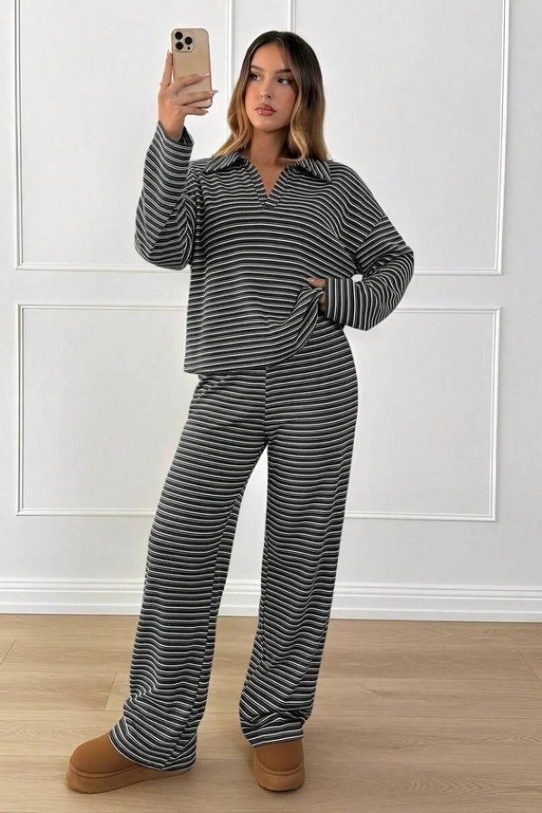A woman takes a mirror selfie wearing the Striped Collared Lounge Set, which features a collared pullover and wide-leg pants in black and white stripes. She pairs the outfit with brown slippers, standing on a light wood floor against a white panel wall.