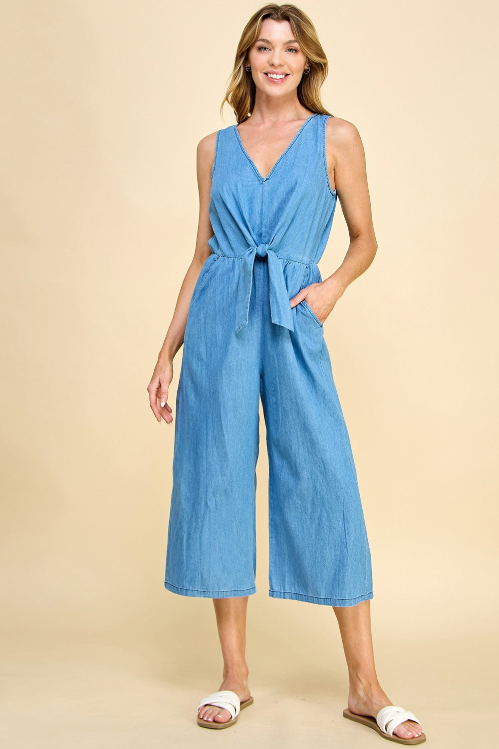 A woman smiles in a Chambray Denim Jumpsuit with a waist tie, relaxed fit, and wide gaucho legs. She poses with one hand in her pocket, wearing white slide sandals against a beige background.