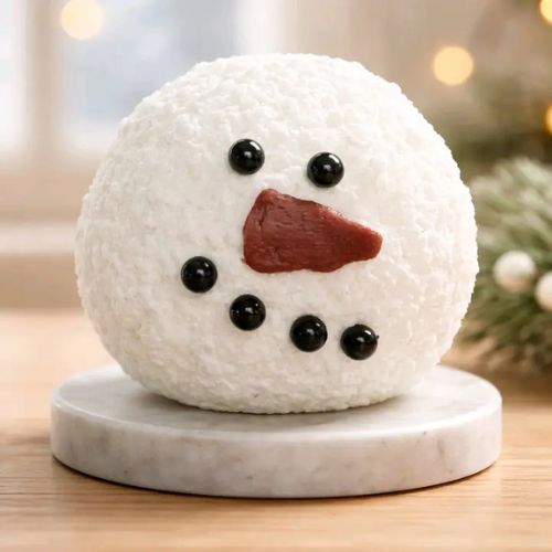 A Snowman Moving Flame Candle, featuring a round shape with a textured white exterior, black details for the eyes, mouth, and buttons, and a bright red nose, is displayed on a marble plate. The scene evokes the look of a charming snowman dessert against a backdrop of blurred holiday lights and festive greenery.