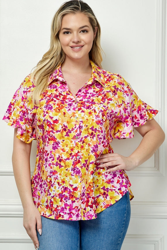 A woman with long blonde hair smiles in a Plus Floral Button Blouse featuring pink, yellow, and orange florals, paired with blue jeans. She stands against a white wall with her hand on her hip.