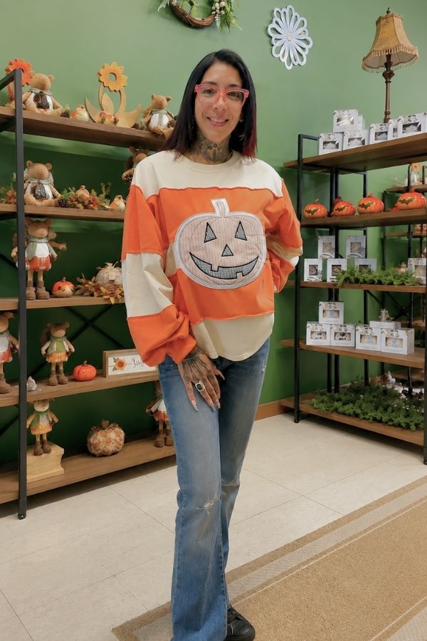 A woman wearing a Gingham Pumpkin Sweatshirt and jeans stands smiling in a store decorated with fall and pumpkin items on shelves, against a green wall.