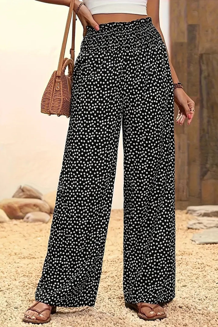 A woman in Polka Dot Wide Leg Pants with an elastic waistband, a white crop top, brown sandals, and a woven handbag stands on a gravel path with decorative stones behind her.