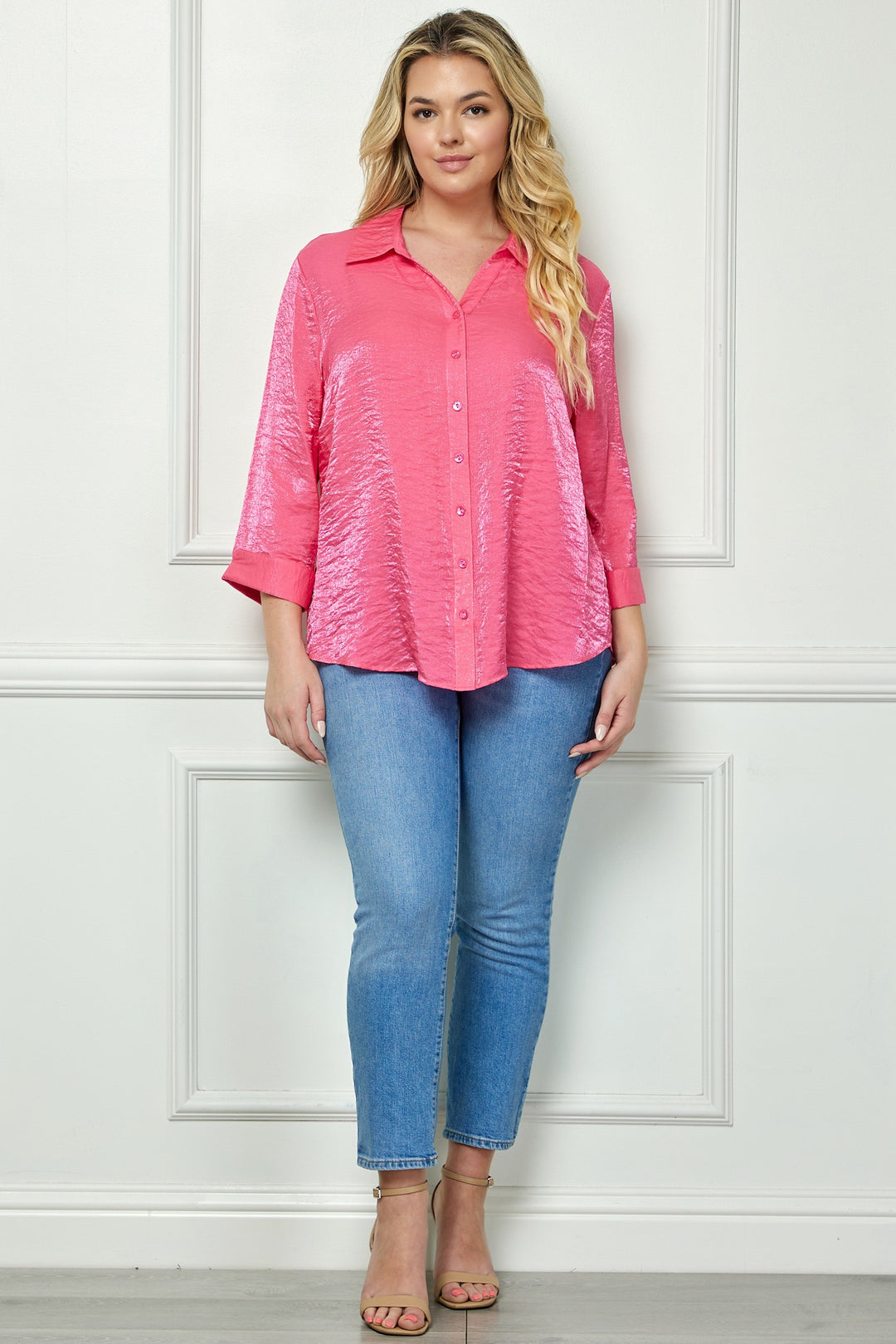 A woman with long wavy blonde hair stands indoors against a white wall, wearing the Plus Shimmer Collar Blouse in pink with three-quarter sleeves, paired with light blue skinny jeans and beige open-toe heels.