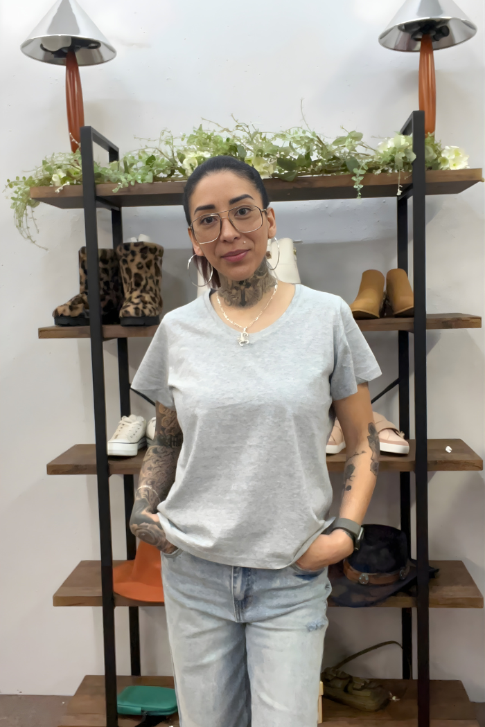 A woman with glasses and tattoos, wearing the Cotton V-Neck Tee and light jeans, stands with her hands in her pockets in front of a shelf displaying shoes, plants, and decorative lamps.
