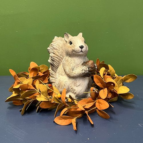 Add a touch of farmhouse charm to your fall décor with the Scamper Squirrel Figurine—a delightful ceramic squirrel holding an acorn, nestled in a ring of orange and yellow leaves, set against a green wall and blue surface.