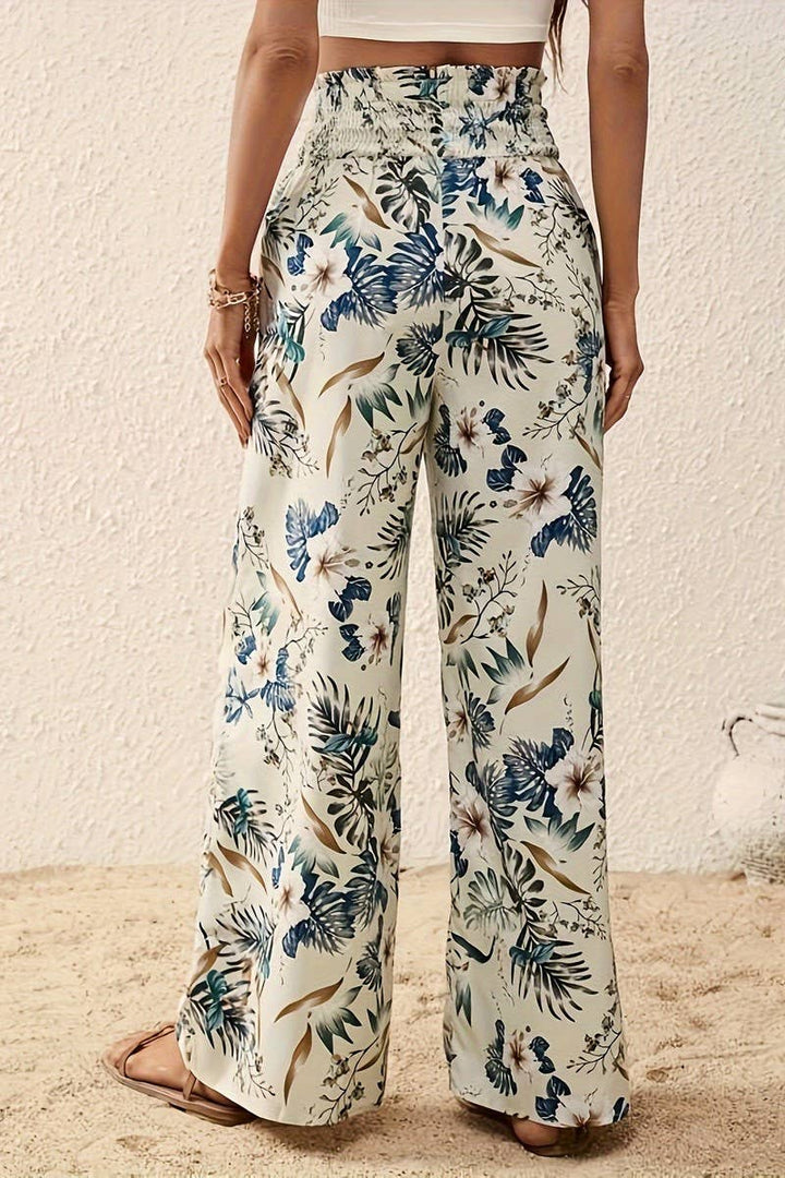Wearing Tropical Wide Leg Pants with a floral print and high-rise elastic waistband, a person stands on sand in front of a textured white wall, pairing them with a white crop top and sandals.