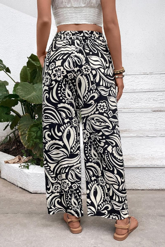 A person stands outdoors with their back turned, wearing Boho Wide Leg Pants featuring a high-rise elastic waist and black and white floral paisley print, paired with a white crop top and sandals. Green plants and white steps are in the background.