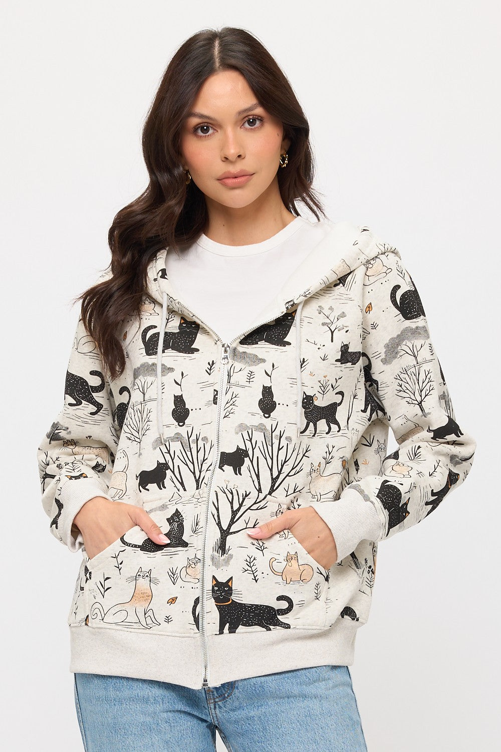 A woman with long brown hair wears the Cat Forest Zip Hoodie, featuring a cat and tree pattern, over a white t-shirt and blue jeans. She stands against a plain white background with her hands in her pockets.