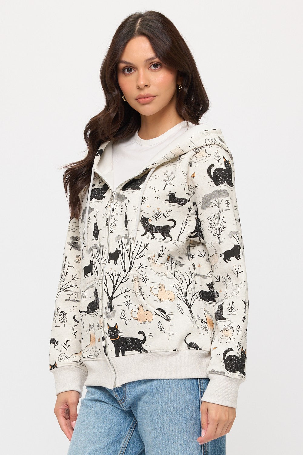 A woman with long brown hair and blue jeans stands against a plain background, modeling the Cat Forest Zip Hoodie—a cozy zip-up featuring a whimsical black and orange cat and forest print—while looking directly at the camera.