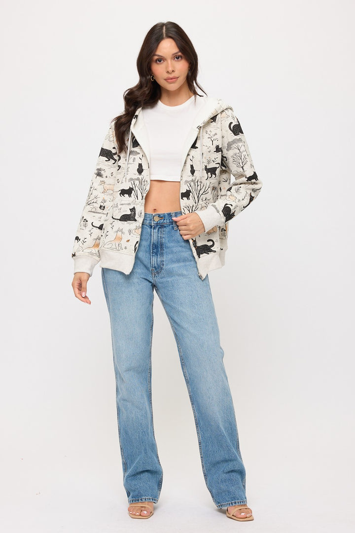 A woman with long, wavy dark hair stands facing forward in a white crop top, light blue high-waisted jeans, beige open-toe heels, and the Cat Forest Zip Hoodie.