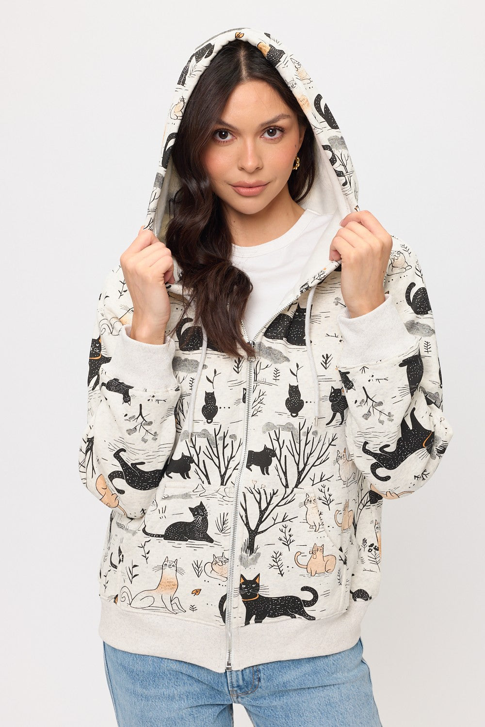 A woman with long dark hair and jeans wears the Cat Forest Zip Hoodie, featuring a playful cat and forest print. She holds the hood with both hands while standing against a plain white background.