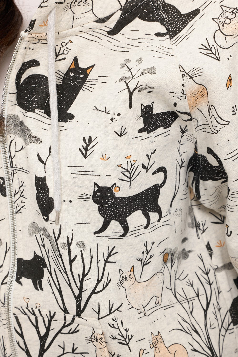 The Cat Forest Zip Hoodie is a cozy cream zip-up featuring playful black and light brown cats posing among trees and shrubs, creating a charming forest scene perfect for any cat lover.