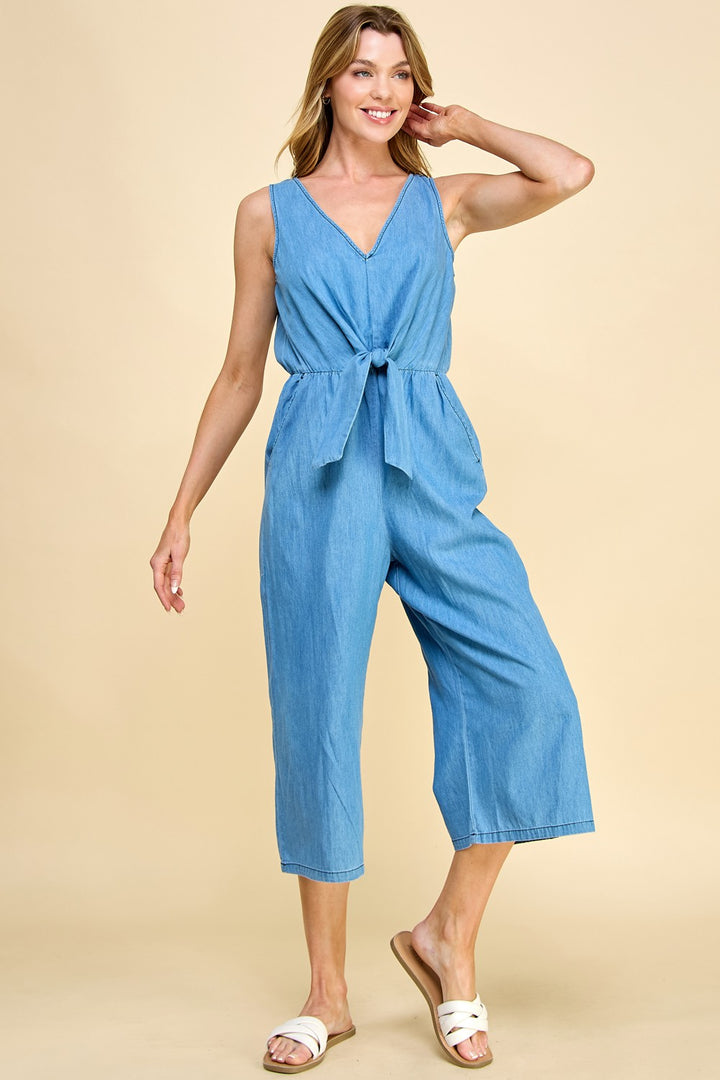 A woman smiles while standing against a beige background, wearing the Chambray Denim Jumpsuit featuring a relaxed fit, front tie, and gaucho leg. She pairs it with white slide sandals.