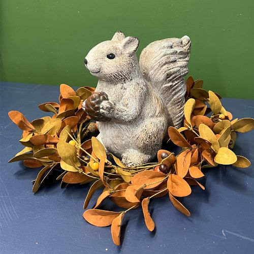 The Scamper Squirrel Figurine features a ceramic squirrel holding an acorn, surrounded by a wreath of orange and yellow leaves on a blue surface with a green background—an adorable accent perfect for fall decorating or adding charm to your farmhouse décor.