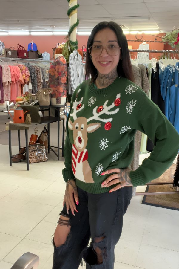 A woman smiles in a store, wearing glasses, black ripped jeans, and the Sequin Reindeer Christmas Sweater. Clothing racks and bags are visible in the background.