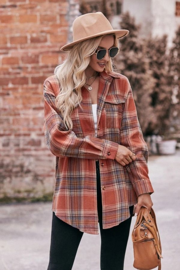 A woman with long wavy blonde hair stands outdoors in front of a brick wall with plants, wearing sunglasses, a beige hat, and the Oversized Plaid Button Shirt in caramel multicolor. She holds a tan handbag.