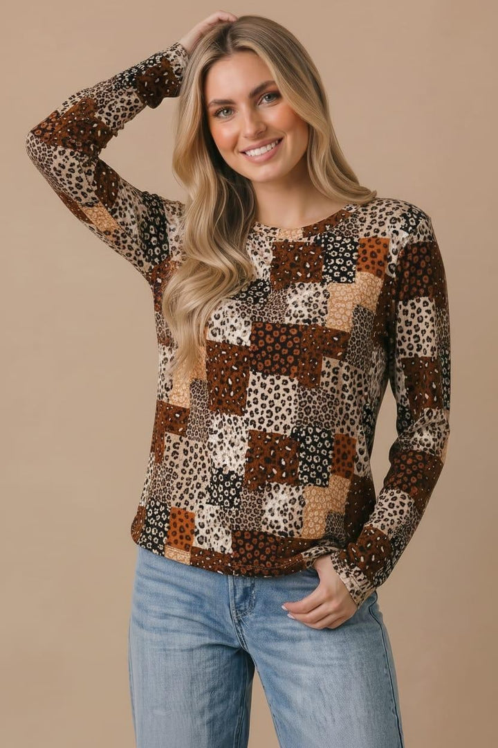 A woman with long blonde hair wears the Plus Animal Patchwork Top—a relaxed fit, long sleeve shirt featuring brown, tan, and animal print patchwork. She smiles while standing against a beige background, with one hand touching her head and the other in her pocket.