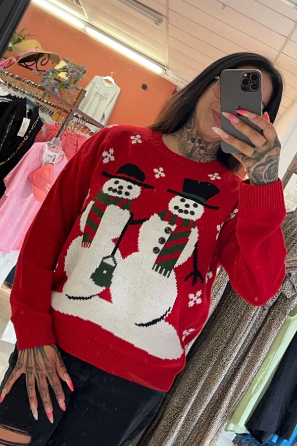 A person takes a mirror selfie wearing the Snowman Christmas Knit Sweater in bright red, which features two snowmen with scarves and hats, along with charming snowflake designs. Clothing racks and a fitting room can be seen in the background.