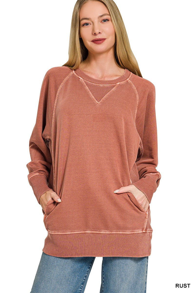 A woman smiles with straight, long hair and her hands in the front pockets, wearing the Pigment Dyed Pullover—a loose, rust-colored French terry sweatshirt with ribbed cuffs and hem—paired with light blue jeans.