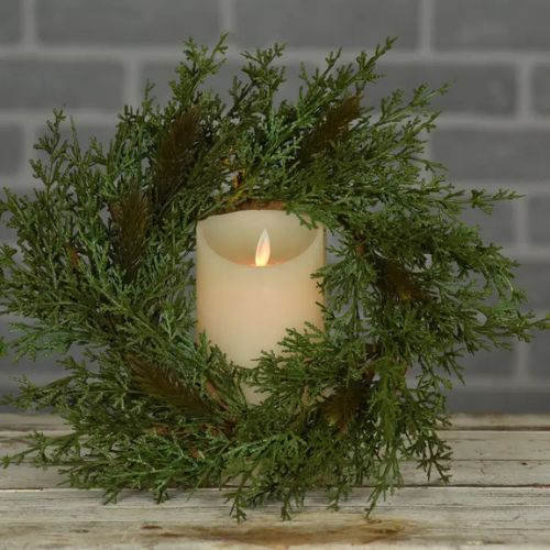 A white candle with a soft flame is nestled in the Rustic Cedar Candle Ring, displayed on a rustic wooden surface with a gray brick wall in the background to create an inviting holiday tabletop arrangement.